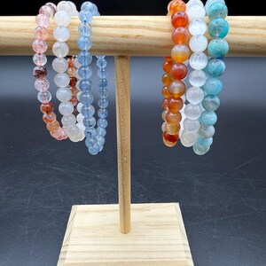 May include: Six elastic bracelets made of round polished gemstone beads in various colors including translucent pink, white, light blue, orange, and teal, displayed on a light-colored wooden T-bar stand against a solid black background.