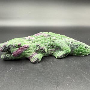 May include: A carved alligator figurine in shades of green and purple. The sculpture is made of a stone material, with a textured surface. The alligator is lying down, with its legs and tail visible. The figurine is a decorative item.