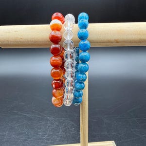 May include: Three beaded bracelets arranged in a row. The left bracelet features smooth, round orange and red carnelian beads. The center bracelet consists of clear, transparent quartz beads. The right bracelet displays round blue apatite beads.
