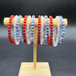 May include: A collection of ten elastic beaded bracelets displayed on a light wood T-bar stand. The bracelets feature round beads in solid red, clear transparent, and mottled blue and white stone patterns, arranged in a repeating sequence.