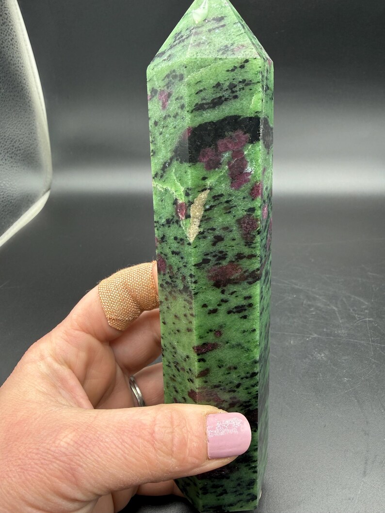 Op de afbeelding: Een groene en zwarte zoisiet kristallen toren met robijn insluitsels. De gepolijste edelsteen heeft een rechthoekige vorm met een puntige top. De kristal wordt in een hand gehouden.