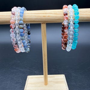 May include: Six beaded bracelets displayed on a wooden T-bar stand. The left side features three bracelets with clear, red-speckled, and blue-toned beads. The right side features three bracelets with dark red, clear, and bright turquoise beads.