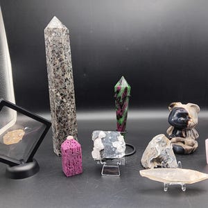 May include: A collection of various crystals and gemstones, including a tall, speckled gray obelisk, a pink hexagonal crystal, and a small bear figurine. Other crystals include a ruby zoisite tower and a clear quartz.