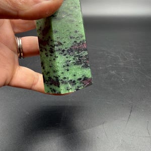 Op de afbeelding: Een groene en zwarte zoisietkristal met rode insluitsels. De gepolijste steen wordt in een hand gehouden en toont zijn natuurlijke patronen en texturen. Het oppervlak van het kristal heeft een mix van groene, zwarte en rode tinten.