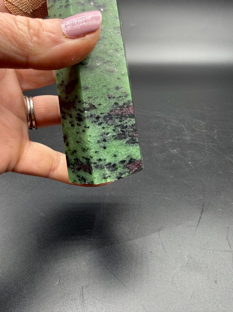 Op de afbeelding: Een groene en zwarte zoisietkristal met rode insluitsels. De gepolijste steen wordt in een hand gehouden en toont zijn natuurlijke patronen en texturen. Het oppervlak van het kristal heeft een mix van groene, zwarte en rode tinten.