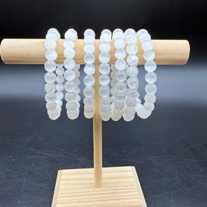 May include: A collection of seven beaded bracelets made from translucent, milky white spherical stones. The bracelets are arranged in a row, hanging from a light-colored wooden T-bar display stand with a square base and a cylindrical horizontal bar.