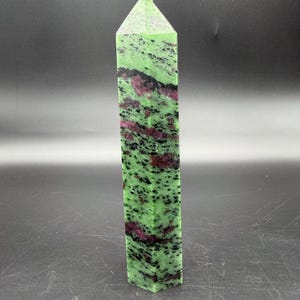 Op de afbeelding: Een hoge, rechthoekige kristallen obelisk. De steen is een levendig groen met zwarte spikkels en vlekken van dieprood. De bovenkant heeft de vorm van een puntige piramide.