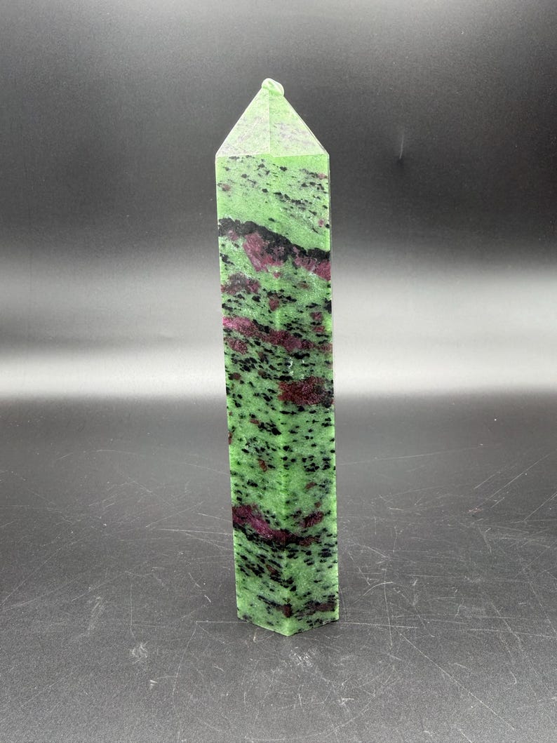 Op de afbeelding: Een hoge, rechthoekige kristallen obelisk. De steen is een levendig groen met zwarte spikkels en vlekken van dieprood. De bovenkant heeft de vorm van een puntige piramide.