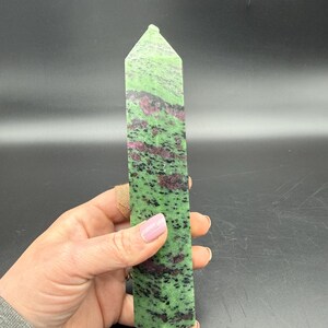 Op de afbeelding: Een groene en rode zoisiet kristallen toren. De gepolijste steen heeft een rechthoekige vorm met een puntige top. De groene kleur is bespikkeld met zwarte en rode insluitsels. De kristal wordt in een hand gehouden.