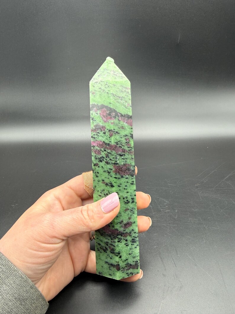 Op de afbeelding: Een groene en rode zoisiet kristallen toren. De gepolijste steen heeft een rechthoekige vorm met een puntige top. De groene kleur is bespikkeld met zwarte en rode insluitsels. De kristal wordt in een hand gehouden.