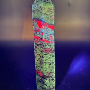 Op de afbeelding: Een hoge, rechthoekige unakiet kristallen obelisk. De steen is voornamelijk groen met rode en zwarte insluitsels. De bovenkant van de obelisk wordt verlicht met een blauw licht. De obelisk is een gepolijst, decoratief item.