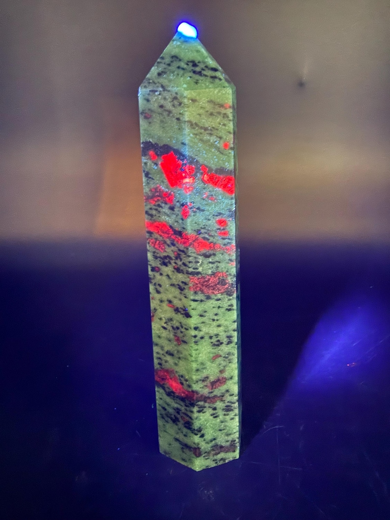 Op de afbeelding: Een hoge, rechthoekige unakiet kristallen obelisk. De steen is voornamelijk groen met rode en zwarte insluitsels. De bovenkant van de obelisk wordt verlicht met een blauw licht. De obelisk is een gepolijst, decoratief item.