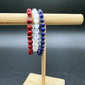 May include: Three beaded stretch bracelets displayed on a wooden T-bar stand. One bracelet features red spherical beads, one consists of translucent white beads, and the third is made of deep blue spherical beads, each arranged in a single strand.