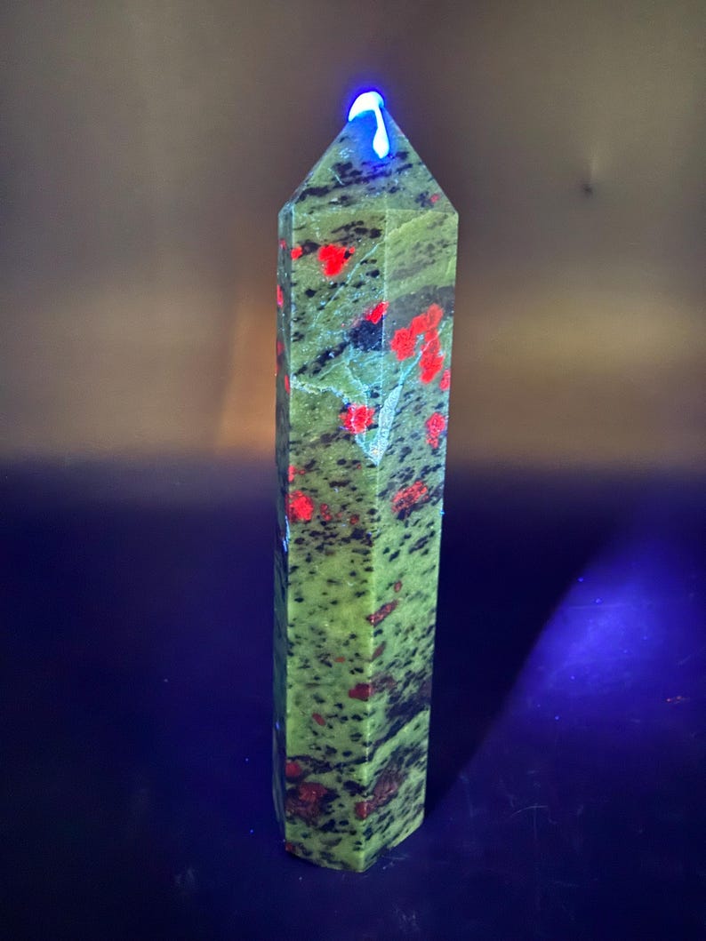 Op de afbeelding: Een hoge, zeshoekige kristallen obelisk. De steen is een gevlekt groen met rode en zwarte insluitsels. De bovenkant van de obelisk gloeit met een helder blauw licht. De kristal is een natuurlijk, decoratief item.
