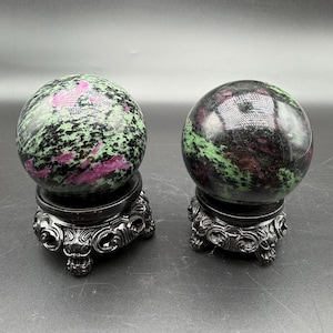 May include: Two polished gemstone spheres, one with a vibrant green and pink pattern, the other with a similar pattern but a darker hue. Each sphere rests on a decorative, dark-colored stand with ornate detailing.