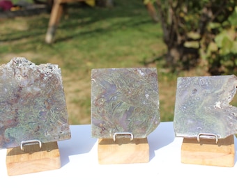 May include: Three polished stone slabs with natural green, brown, and white patterns. Each slab is mounted on a small wooden stand with a metal bracket. The slabs have irregular edges and varying shapes, displayed outdoors.