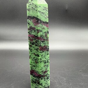 Op de afbeelding: Een hoge, rechthoekige kristallen obelisk. De steen is een levendig groen met zwarte spikkels en vlekken van dieprood. De bovenkant heeft een puntige, facetvorm.