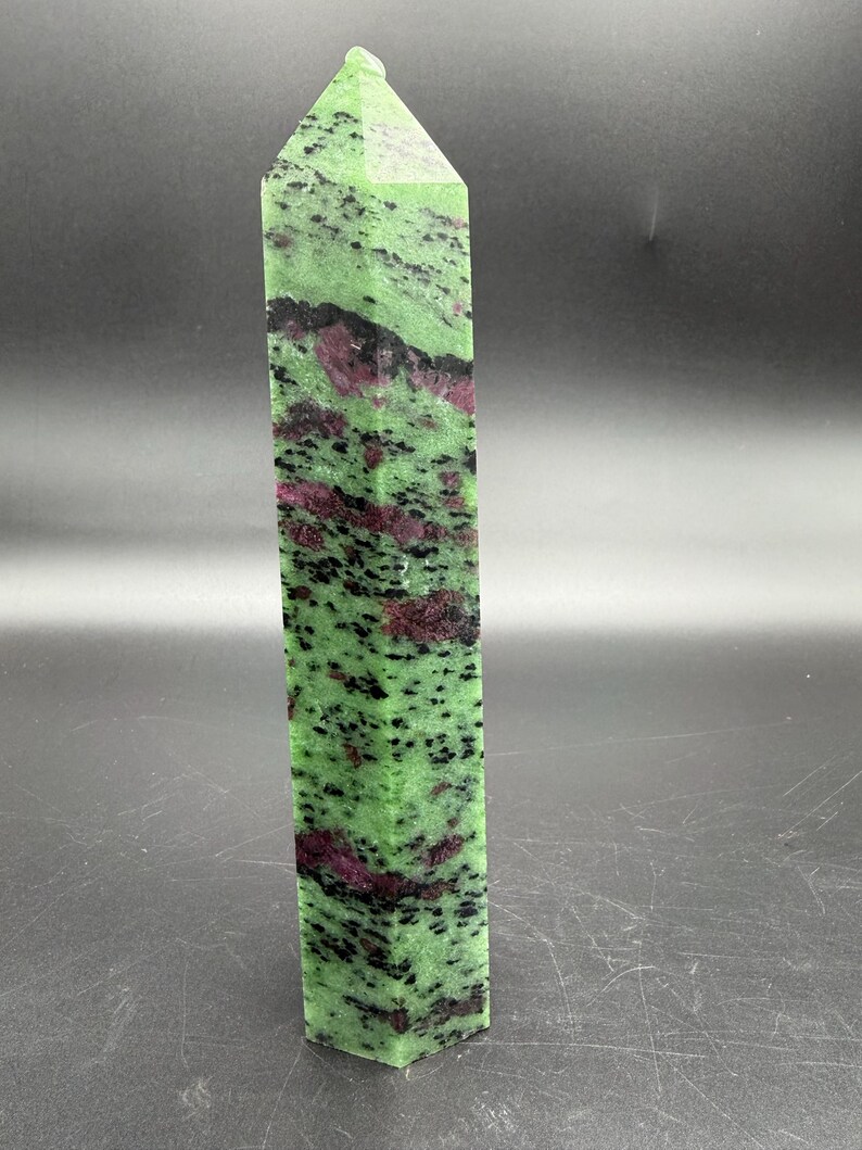 Op de afbeelding: Een hoge, rechthoekige kristallen obelisk. De steen is een levendig groen met zwarte spikkels en vlekken van dieprood. De bovenkant heeft een puntige, facetvorm.