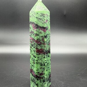 May include: A tall, rectangular zoisite crystal with a pointed top. The stone is primarily green with dark red and black inclusions. The crystal is standing upright against a dark background.