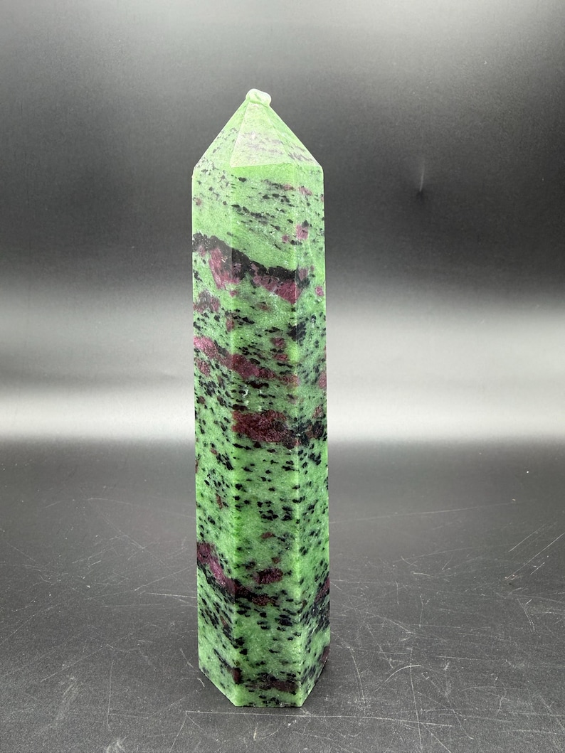 Op de afbeelding: Een lange, rechthoekige zoisietkristal met een puntige top. De steen is voornamelijk groen met donkerrode en zwarte insluitsels. Het kristal staat rechtop tegen een donkere achtergrond.