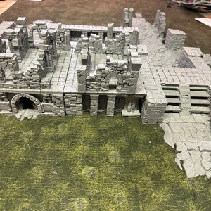 Op de afbeelding: Een grijs, gedetailleerd, 3D-geprint model van een verwoeste tempel of gebouw. Het model bestaat uit meerdere onderdelen en heeft bogen, kolommen en een trap. Het model staat op een groene ondergrond.