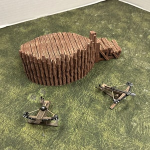 May include: Two wooden catapults with black metal parts on a green grassy surface. The catapults are made of wood and have a black metal arm and a black metal projectile. The catapults are on a green grassy surface.