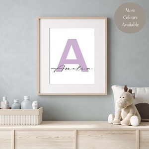 May include: A framed print featuring a large, pale purple letter A with the name Amelia written in cursive underneath. The print is on a white background and is framed in a light wood frame.