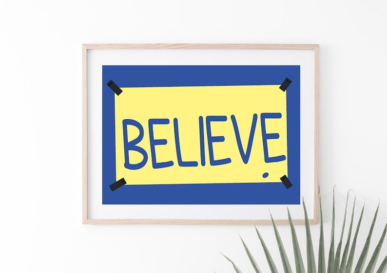 Puede incluir: Una impresi&oacute;n enmarcada con fondo amarillo y borde azul. La palabra "BELIEVE" est&aacute; escrita en azul sobre el fondo amarillo.