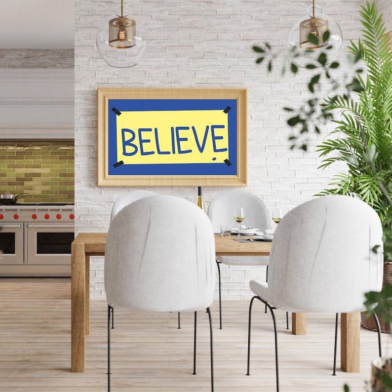 Puede incluir: Un p&oacute;ster enmarcado con fondo amarillo y borde azul, con la palabra "BELIEVE" en texto negro. El p&oacute;ster est&aacute; colgado en una pared de ladrillo blanco sobre una mesa de comedor con dos sillas blancas.