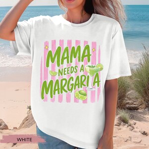 May include: White t-shirt with the text "MAMA NEEDS A MARGARITA" in green, with a pink striped background. The design includes a margarita glass and lime slices. The shirt is a casual, relaxed fit.