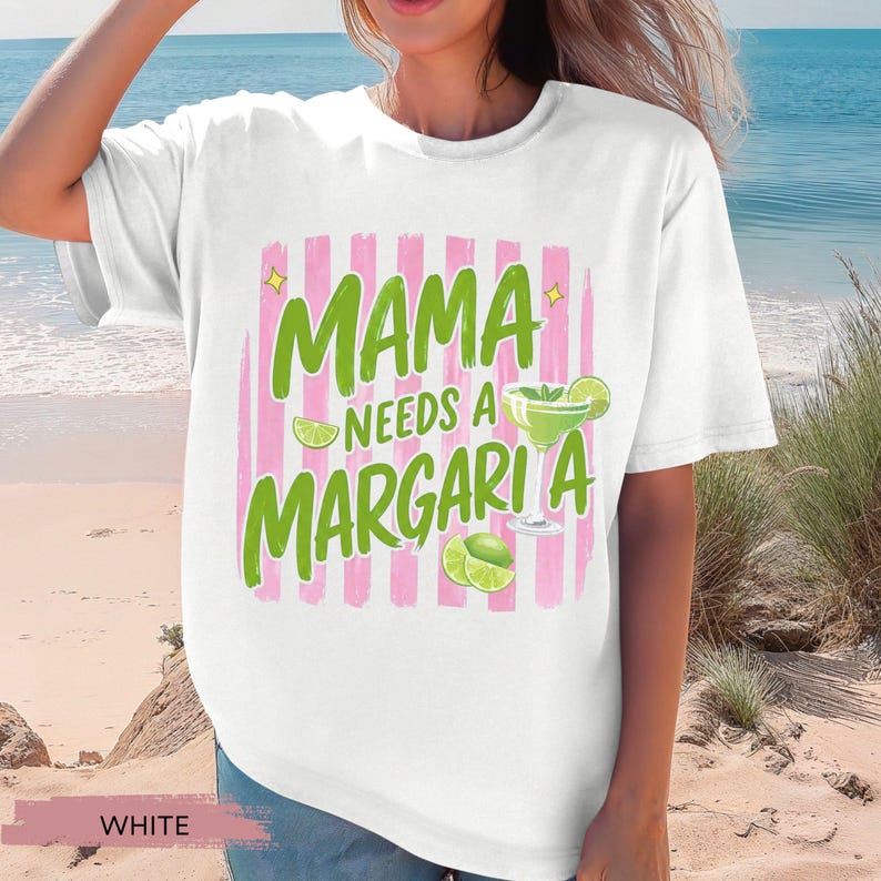 May include: White t-shirt with the text "MAMA NEEDS A MARGARITA" in green, with a pink striped background. The design includes a margarita glass and lime slices. The shirt is a casual, relaxed fit.