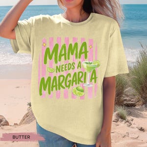 May include: Butter-colored t-shirt with the text "MAMA NEEDS A MARGARITA" in green, with pink vertical stripes. A margarita glass and lime slices are also featured in the design. The shirt is a casual, relaxed fit.