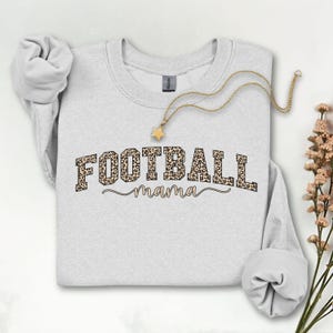 May include: A light gray sweatshirt with the words "FOOTBALL mama" in a leopard print design. A gold necklace with a star pendant is draped across the sweatshirt. The sleeves are rolled up.