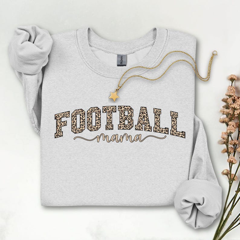 May include: A light gray sweatshirt with the words "FOOTBALL mama" in a leopard print design. A gold necklace with a star pendant is draped across the sweatshirt. The sleeves are rolled up.