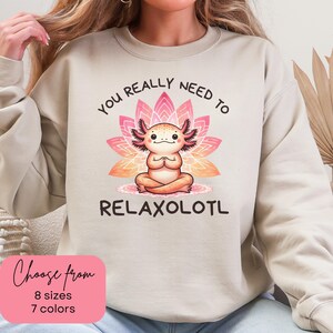 May include: Beige sweatshirt with a graphic of a cartoon axolotl meditating in front of a pink lotus flower. The text reads "YOU REALLY NEED TO RELAXOLOTL". A pink rectangle at the bottom left says "Choose from 8 sizes 7 colors".