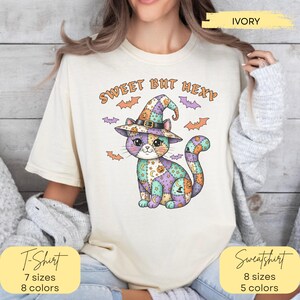 May include: An ivory t-shirt featuring a graphic of a patchwork cat wearing a witch hat, surrounded by small bats. The text "SWEET BUT HEXY" is printed above the cat. The design includes orange, purple, and teal colors on the cat's body.