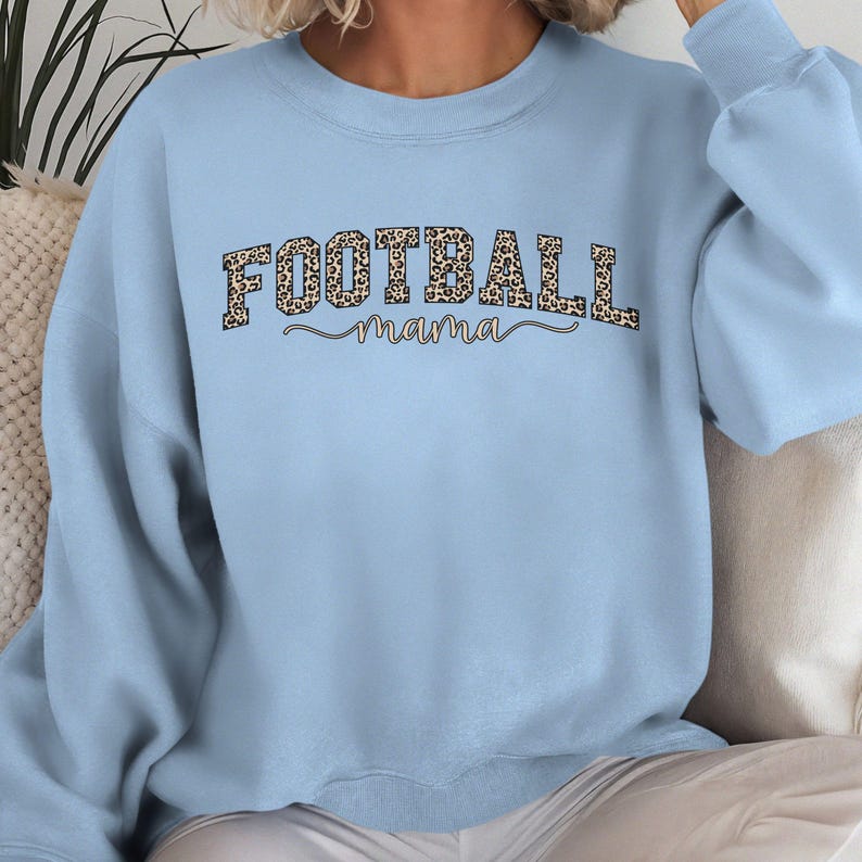 May include: A light blue sweatshirt with the words "FOOTBALL mama" printed across the front. The word "FOOTBALL" is in a leopard print pattern, and "mama" is in a cursive font.