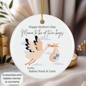 May include: White ceramic ornament with a stork carrying a baby in a bag. The ornament says "Happy Mother's Day Mama to be of twin boys" and "Love, Babies Noah & Liam".