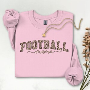May include: A light pink sweatshirt with the text "FOOTBALL mama" in a leopard print design. A gold-colored necklace with a star pendant is draped across the sweatshirt. The sweatshirt's sleeves are folded. A sprig of dried flowers is in the background.