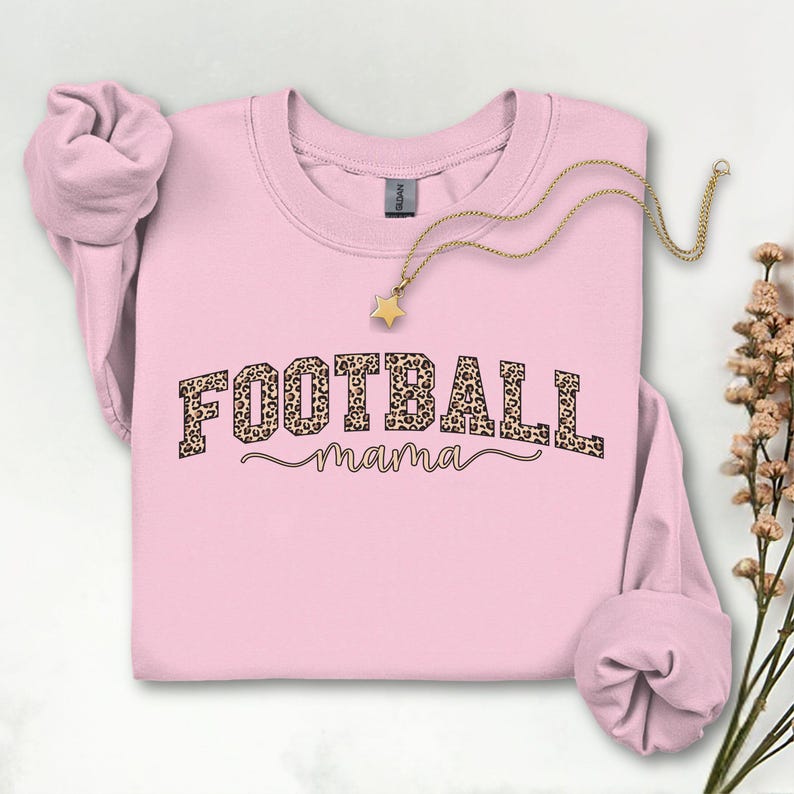 May include: A light pink sweatshirt with the text "FOOTBALL mama" in a leopard print design. A gold-colored necklace with a star pendant is draped across the sweatshirt. The sweatshirt's sleeves are folded. A sprig of dried flowers is in the background.