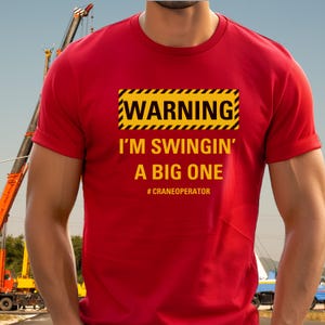 May include: Red t-shirt with a yellow warning sign that says "WARNING I'M SWINGIN' A BIG ONE #CRANEOOPERATOR"
