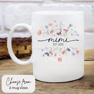 May include: White ceramic mug with a floral design in shades of pink, blue, and green. The mug has the text "mimi est. 2025" printed on it.