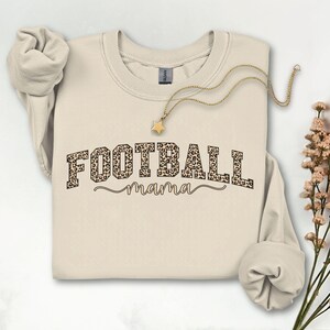 May include: A cream-colored sweatshirt with the words "FOOTBALL mama" printed on the front. The word "FOOTBALL" is in a bold, leopard print font, and "mama" is in a cursive font. A gold necklace with a star pendant is draped across the sweatshirt.
