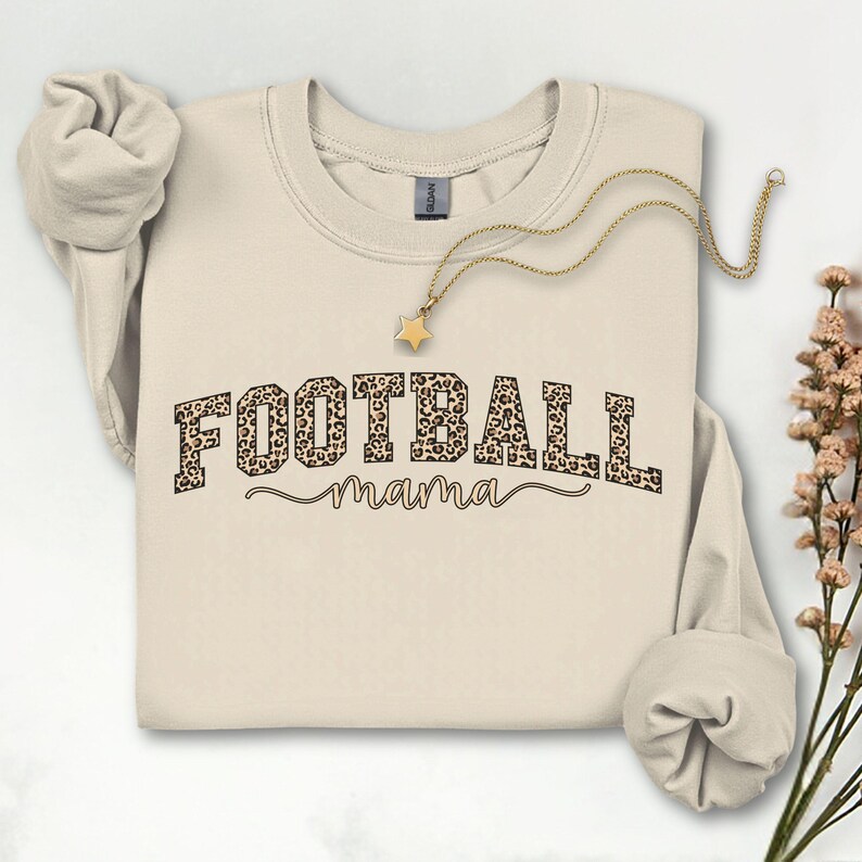 May include: A cream-colored sweatshirt with the words "FOOTBALL mama" printed on the front. The word "FOOTBALL" is in a bold, leopard print font, and "mama" is in a cursive font. A gold necklace with a star pendant is draped across the sweatshirt.