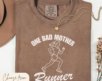 May include: A brown t-shirt with the text "ONE BAD MOTHER RUNNER" in white. A white graphic of a running person is below the text. A gold star pendant necklace is on the shirt. The shirt is available in 7 sizes and 7 colors.