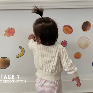 May include: A young child is standing in front of a white wall with colorful fruit stickers on it. The text "STAGE 1 OBJECT RECOGNITION" is printed below the stickers.