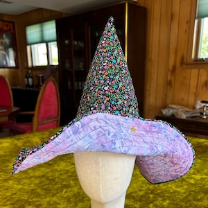 May include: A quilted witch hat featuring a tall, pointed crown with a black fabric base covered in a colorful floral pattern. The wide, floppy brim is lined with a light purple fabric displaying a marbled, wavy texture and a small gold star.