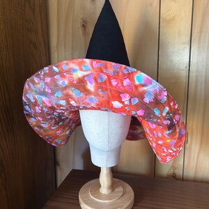May include: A pointed witch hat featuring a solid black conical crown and a wide, quilted brim. The brim displays a vibrant, multi-colored batik-style pattern with splashes of orange, red, purple, and bright blue across the fabric surface.