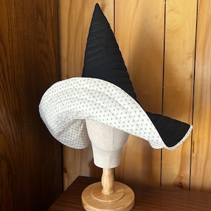 May include: A tall, pointed witch hat featuring a quilted black exterior and a wide, floppy brim. The underside of the brim is white and decorated with a repeating pattern of small, gold-colored stars. The hat is displayed on a neutral mannequin head.
