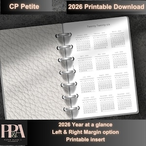 May include: A white 2026 printable planner insert with a textured cover. The calendar pages are open, displaying monthly calendars. The text "Twenty Twenty-six" and "2026 Year at a glance" are visible. The planner is held together with silver rings.